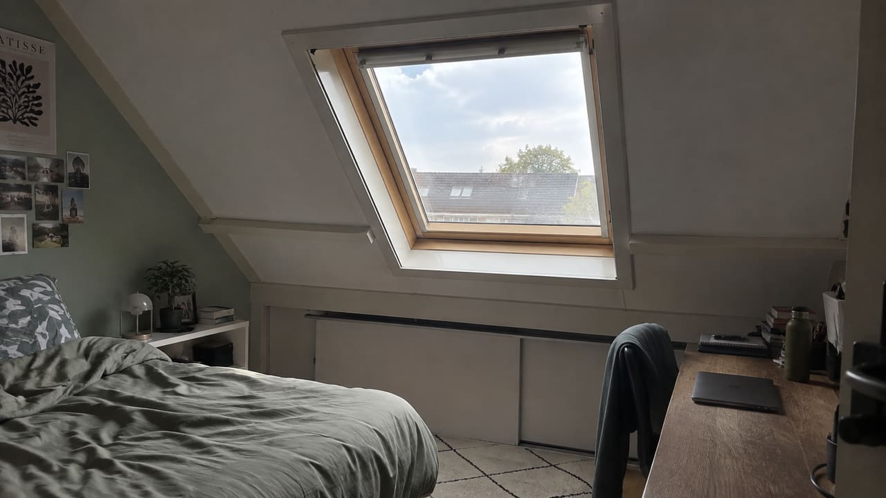Houten dakraam in de schuine dakwand van een zolderkamer met een bed en bureau.