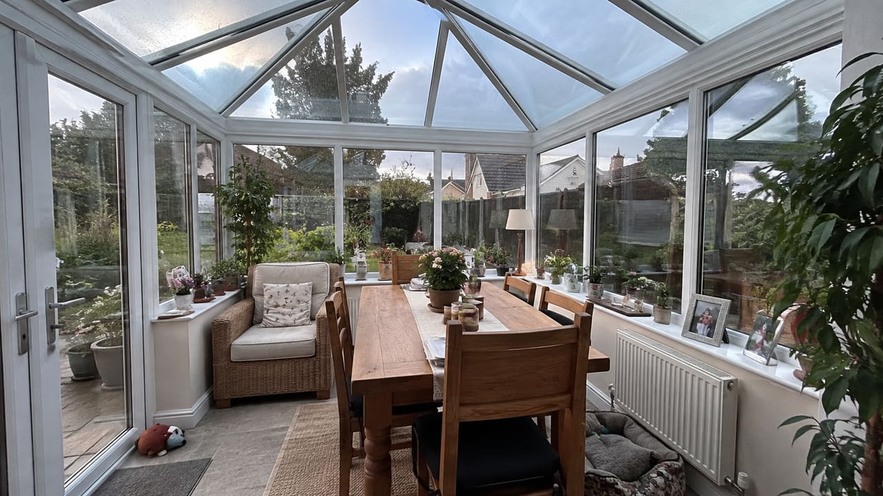 Serre met een glazen dak, grote zijramen en een houten eettafel.